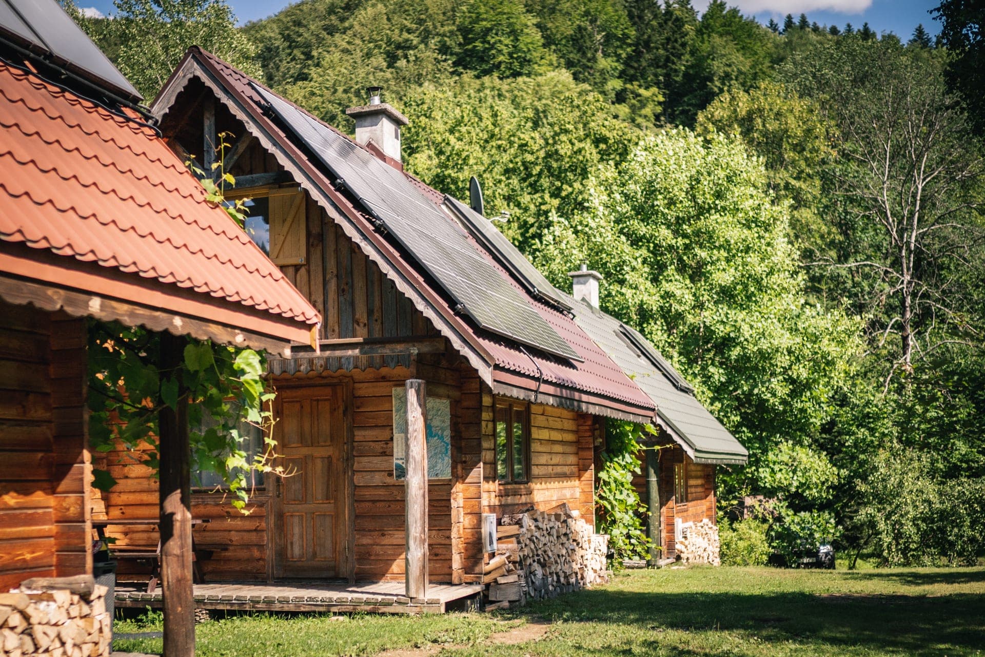 Rząd trzech drewnianych domków na słonecznej łące, za nimi zalesione wzgórze